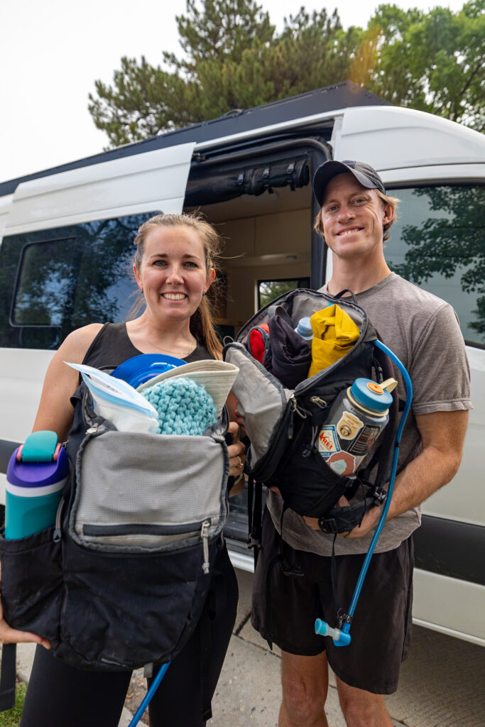 Jen and Elliot are holding their hiking day packs filled with everything they take on hikes from water, spare clothing, sunscreen, first aid kit, bathroom essentials and more.
