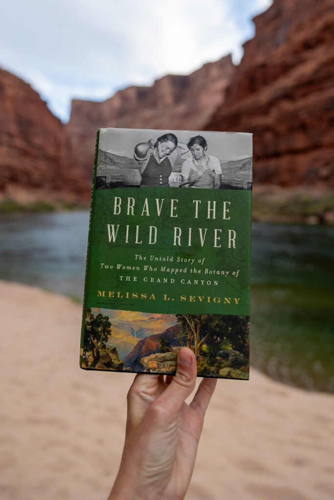 Jen's hand holding "Brave the Wild River" by Melissa L. Sevigny at the Colorado River in the Grand Canyon, with red canyon walls and blue sky in background
