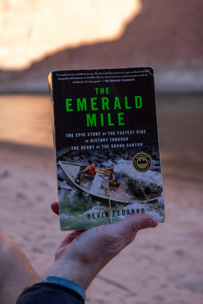 Elliot's hand holding The Emerald Mile book by Kevin Fedarko against a Grand Canyon backdrop at sunset, showing the book's cover with green title text and an image of boatmen navigating rapids