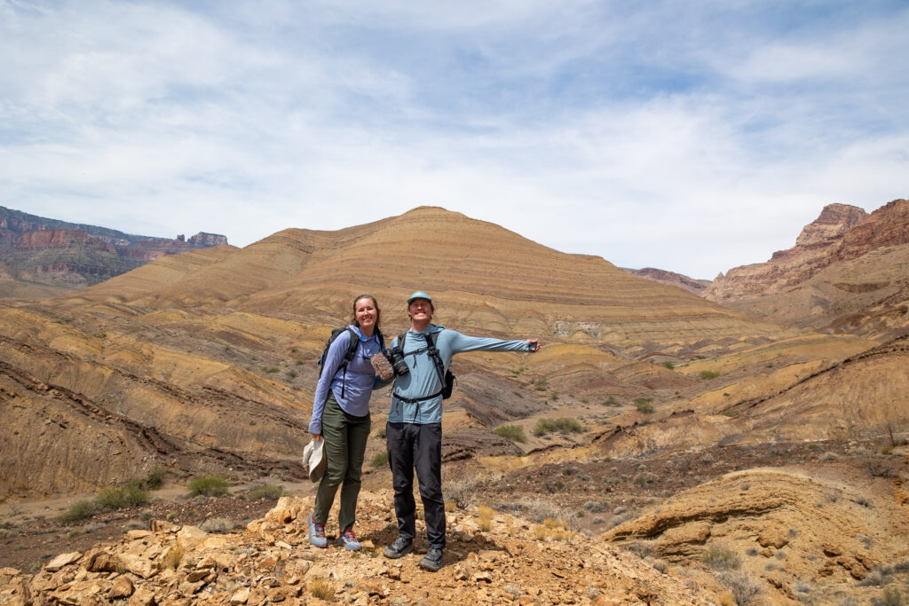 Two hikers in backpacks stand on a rocky outcrop in a vast canyon landscape with tan and rust-colored rock formations under a partly cloudy sky