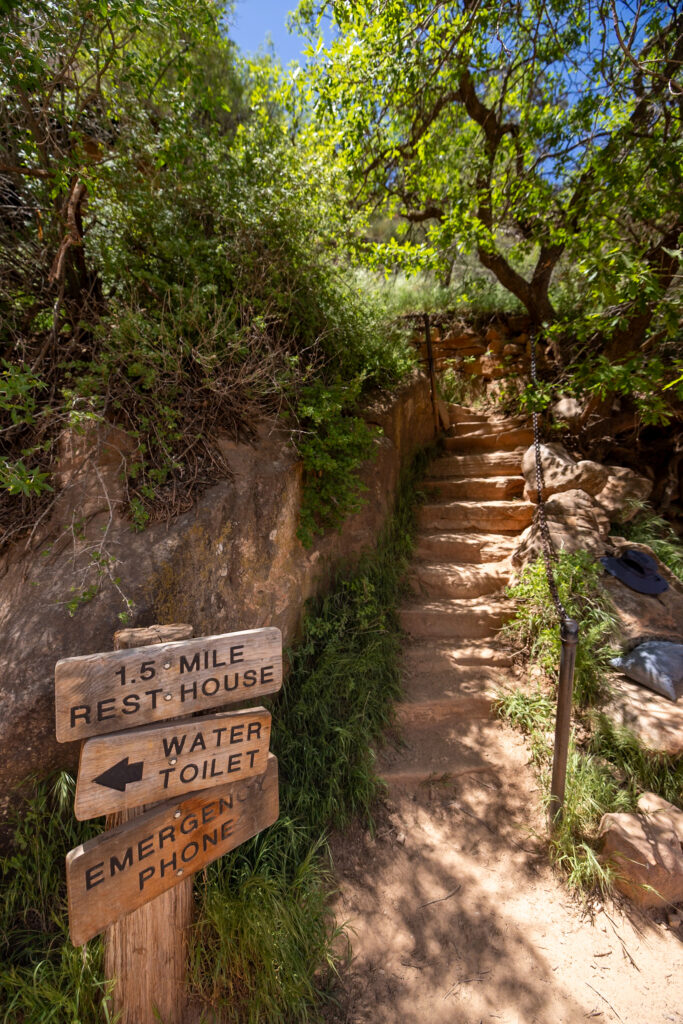 Wooden trail signs for 1.5 Mile Rest House, water, toilet, and emergency phone marking the shaded Bright Angel Trail with stone steps ascending through vegetation