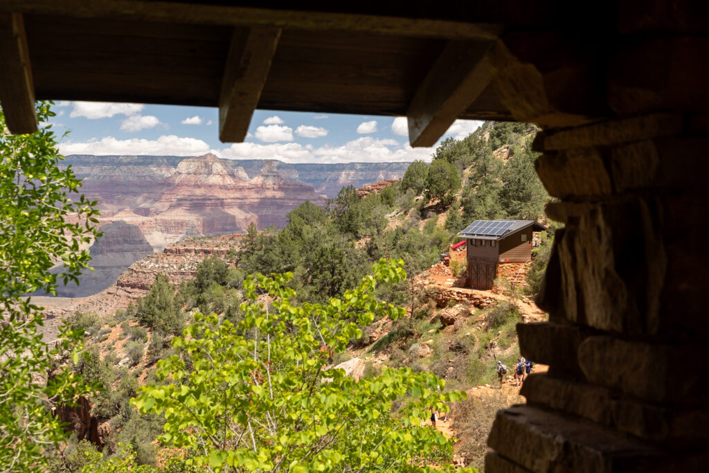 View from inside the 1.5-Mile Resthouse shelter showing canyon landscape with layered rock formations, green vegetation, and a bathroom structure on the trail below