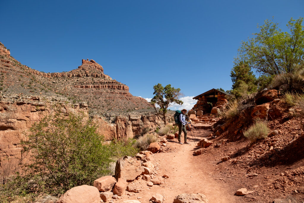 Woman resting at the 3 Mile Resthouse shelter on the Bright Angel Trail, surrounded by red canyon walls and desert vegetation