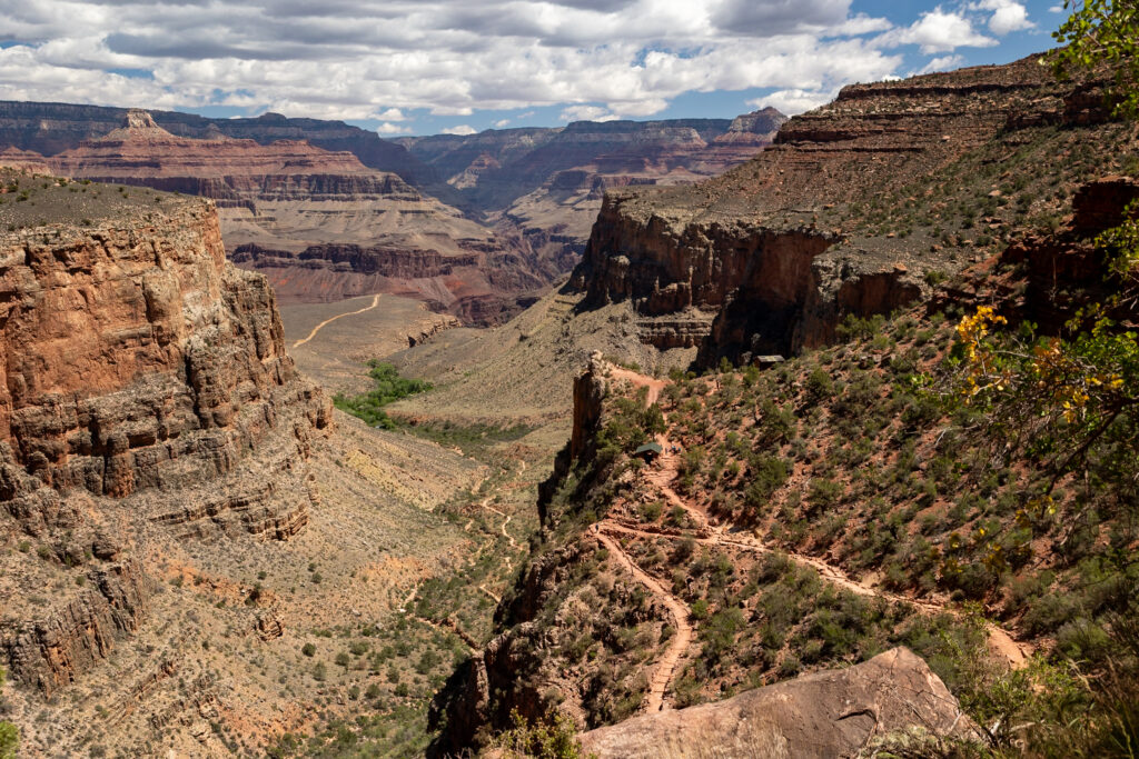 Canyon vista showing layered red rock formations, distant plateau, winding trail through vegetation, and Colorado River visible in the valley below