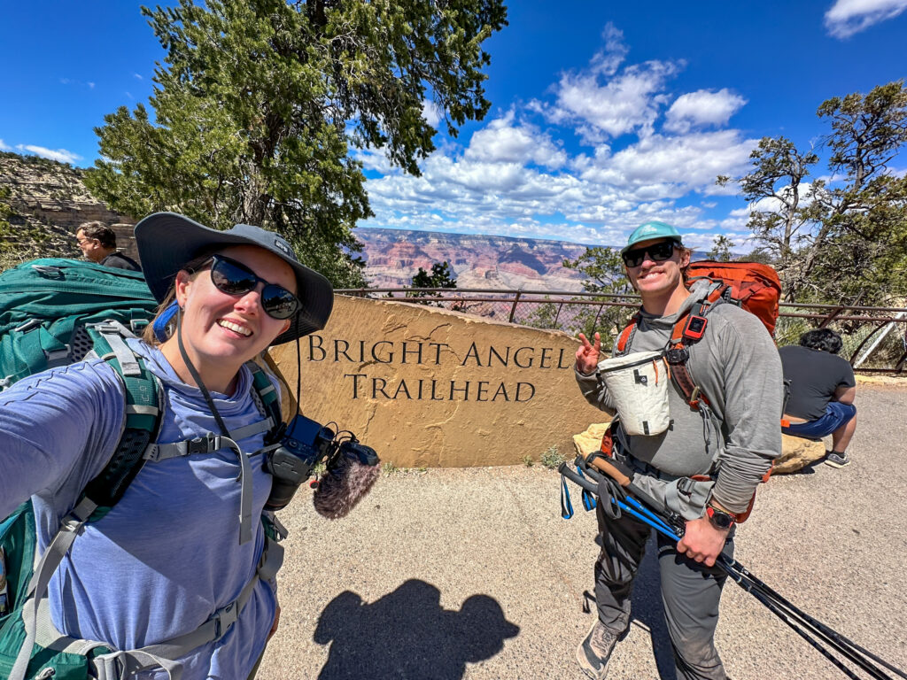 Two hikers posing at the Bright Angel Trailhead sign with the Grand Canyon visible in the background