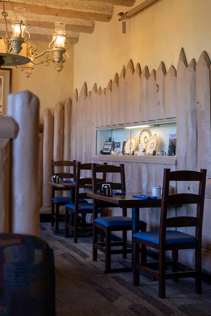 Dining area in Harvey House Cafe at Bright Angel Lodge, featuring wooden tables with blue chairs, decorative white picket fence wall, and vintage brass lighting fixtures