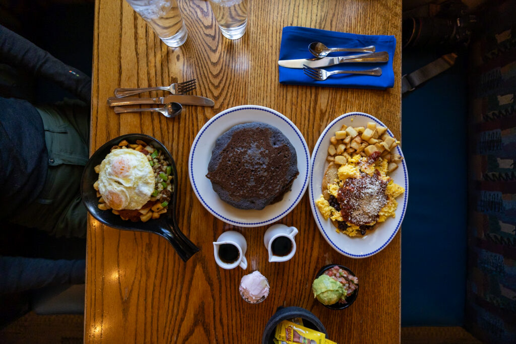 Three breakfast dishes on a wooden table: a skillet with hash browns and fried eggs, a plate of huevos rancheros with chocolate dusting, and prickly pear blue corn cakes with powdered sugar
