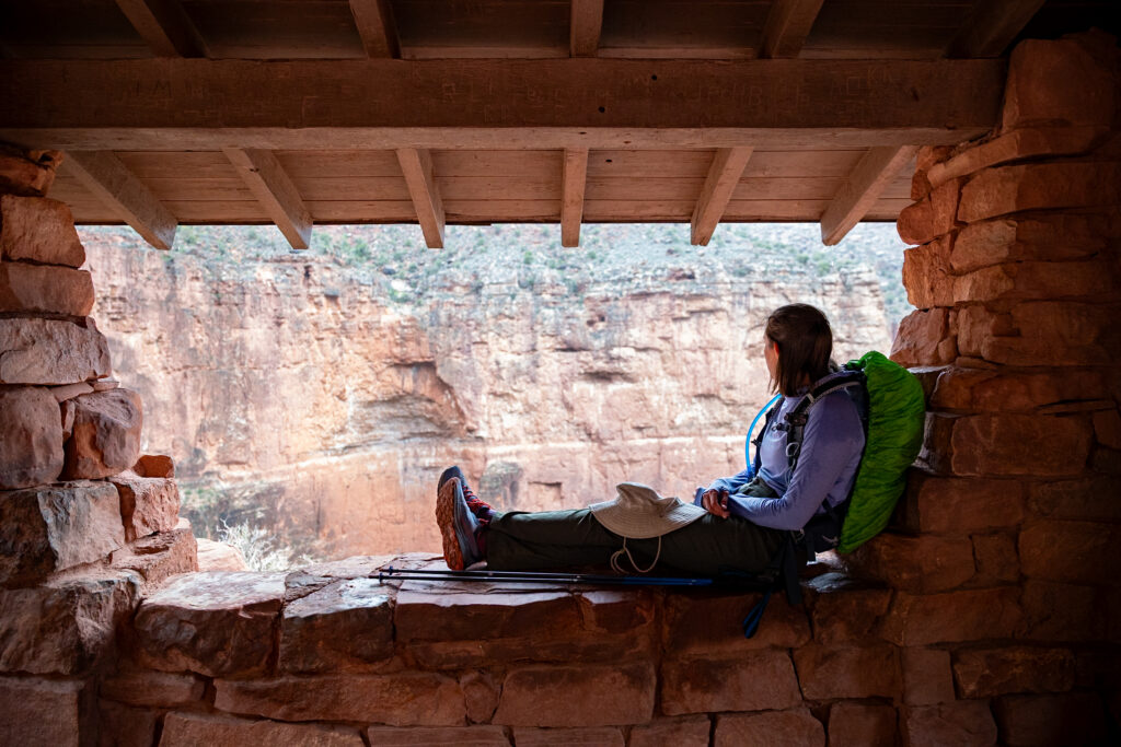 Hiker resting in shaded stone shelter at 3-Mile Resthouse on Bright Angel Trail, with expansive canyon views visible beyond