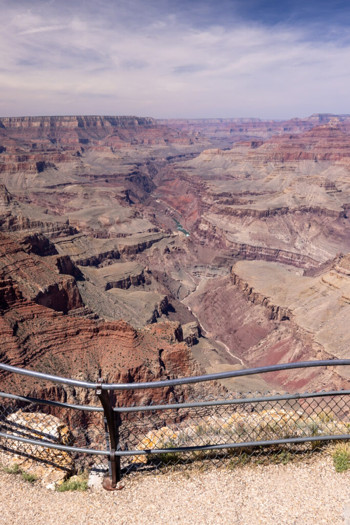 Panoramic view of the Grand Canyon from Lipan Point, showing layered rock formations, the Colorado River winding through the canyon, and distant San Francisco Peaks under a partly cloudy sky