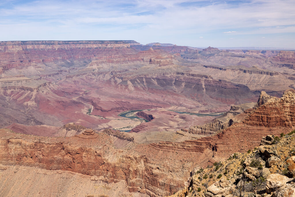 Expansive view of the Grand Canyon from Lipan Point showing layered red rock formations, the Colorado River winding through the canyon floor, and distant plateaus under a partly cloudy sky