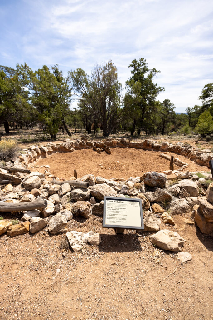 Tusayan Ruins, ancient stone structures arranged in a circle with an interpretive sign, surrounded by desert vegetation and trees along Desert View Drive