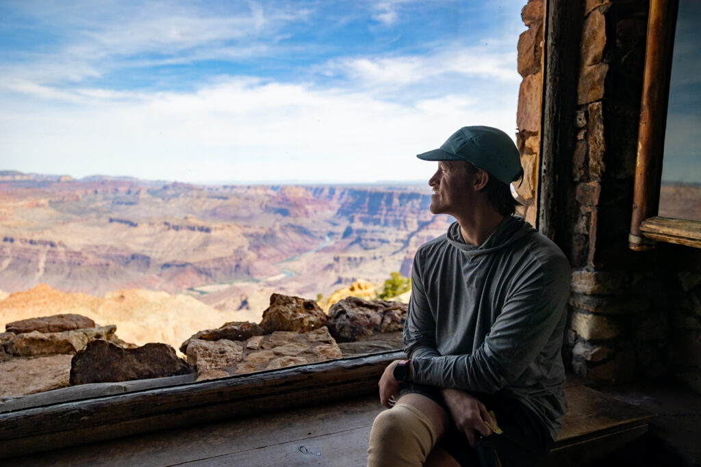 Man sitting on wooden bench at Desert View Watchtower overlooking expansive Grand Canyon landscape with layered rock formations and Colorado River visible below. This is one of the best things to do in the Grand Canyon on the South Rim.