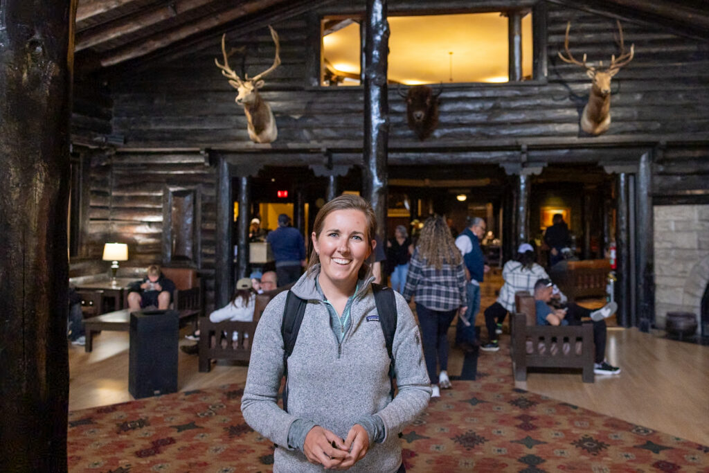 Jen smiling in El Tovar Hotel's rustic log lobby with mounted deer heads and other guests in the background