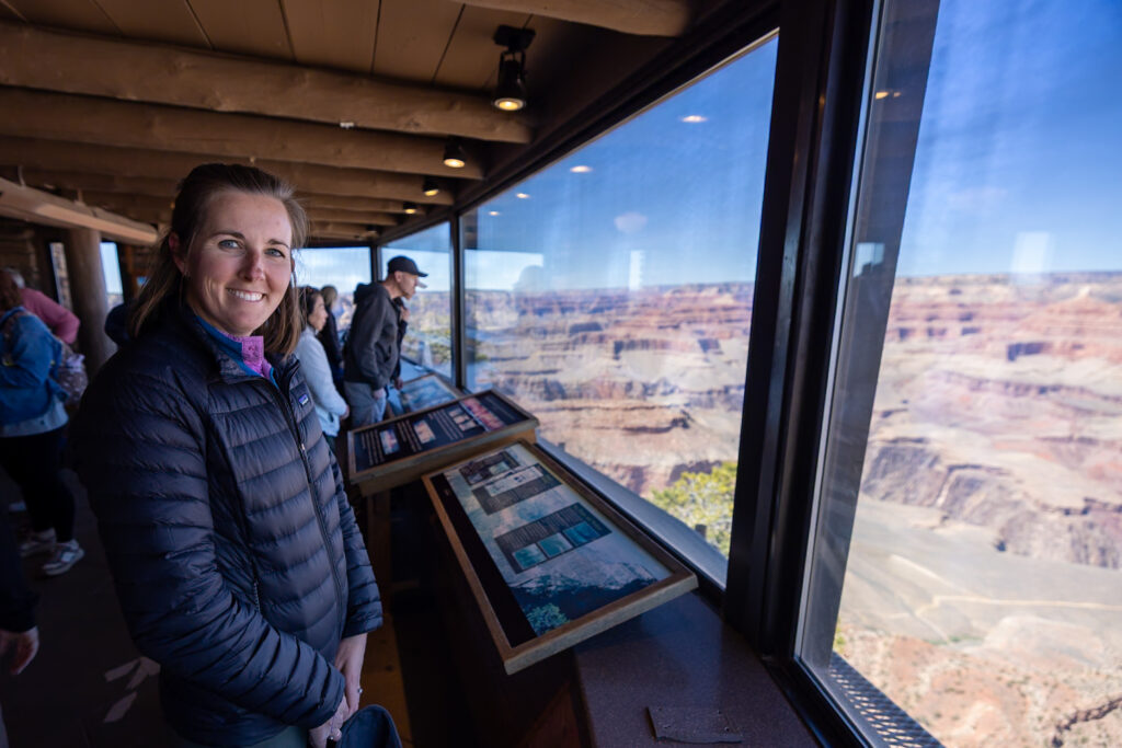Woman smiling at the camera inside the Yavapai Geology Museum, with panoramic windows showing expansive Grand Canyon views and geological display exhibits in the foreground. Visiting the Geology Museum is one of the best things to do at the South Rim of the Grand Canyon.