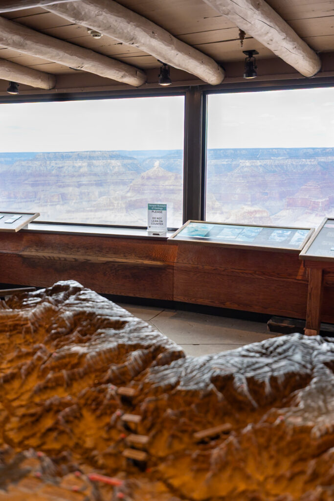 Interior of the Yavapai Geology Museum showing a large topographical relief map in the foreground with panoramic windows framing expansive canyon views beyond. Visiting the Geology Museum is one of the best things to do at the South Rim of the Grand Canyon.