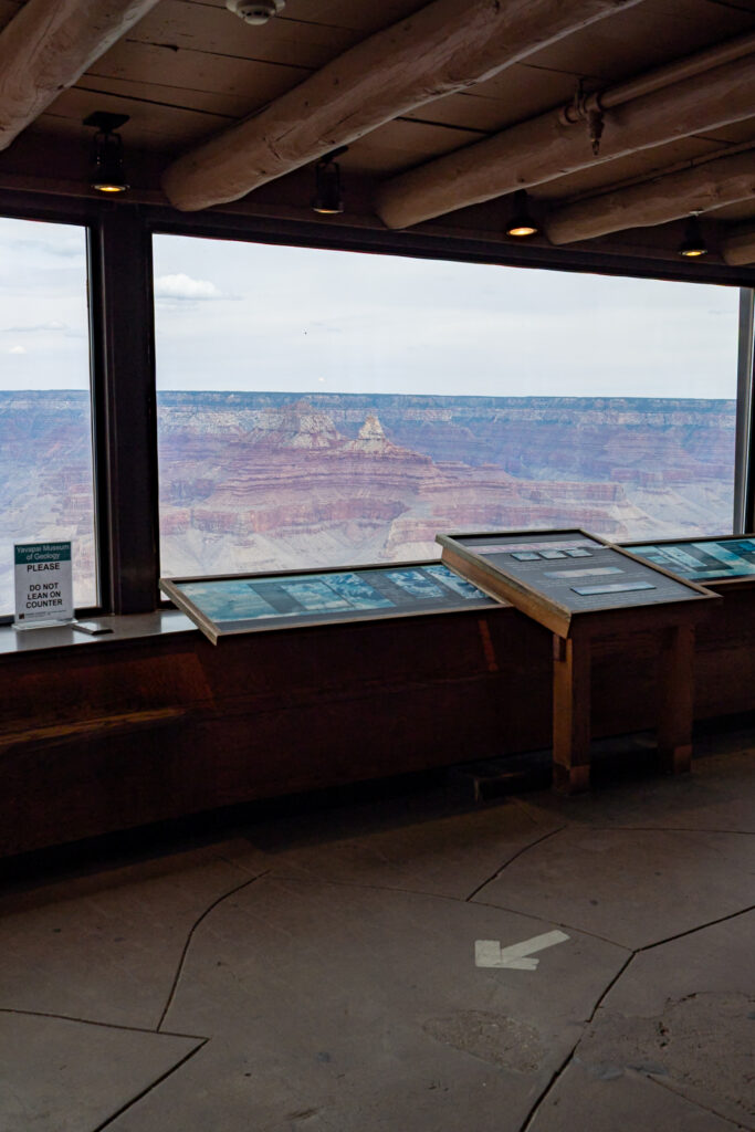 Interior of the Yavapai Geology Museum with large windows framing layered canyon walls and rock formations, educational display tables in foreground