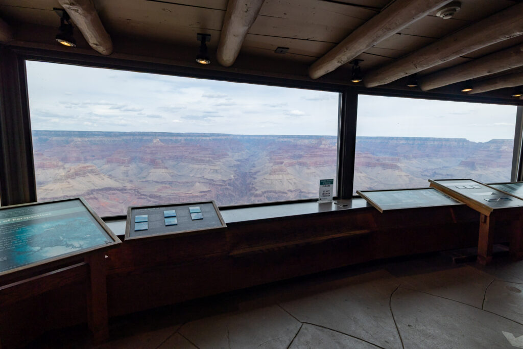 Interior of the Yavapai Geology Museum with large windows framing expansive views of the layered rock formations and canyons of the Grand Canyon, with interpretive displays in the foreground