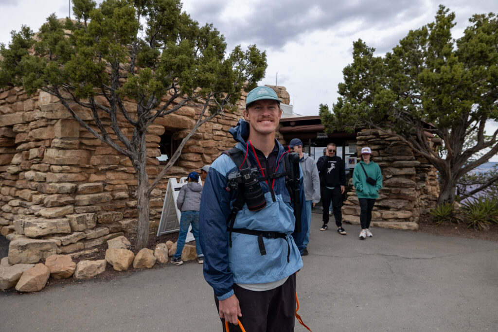 Elliot standing outside the Yavapai Geology Museum, a stone building surrounded by juniper trees, wearing a blue jacket and cap with a camera. Visiting the Geology Museum is one of the best things to do at the South Rim of the Grand Canyon.