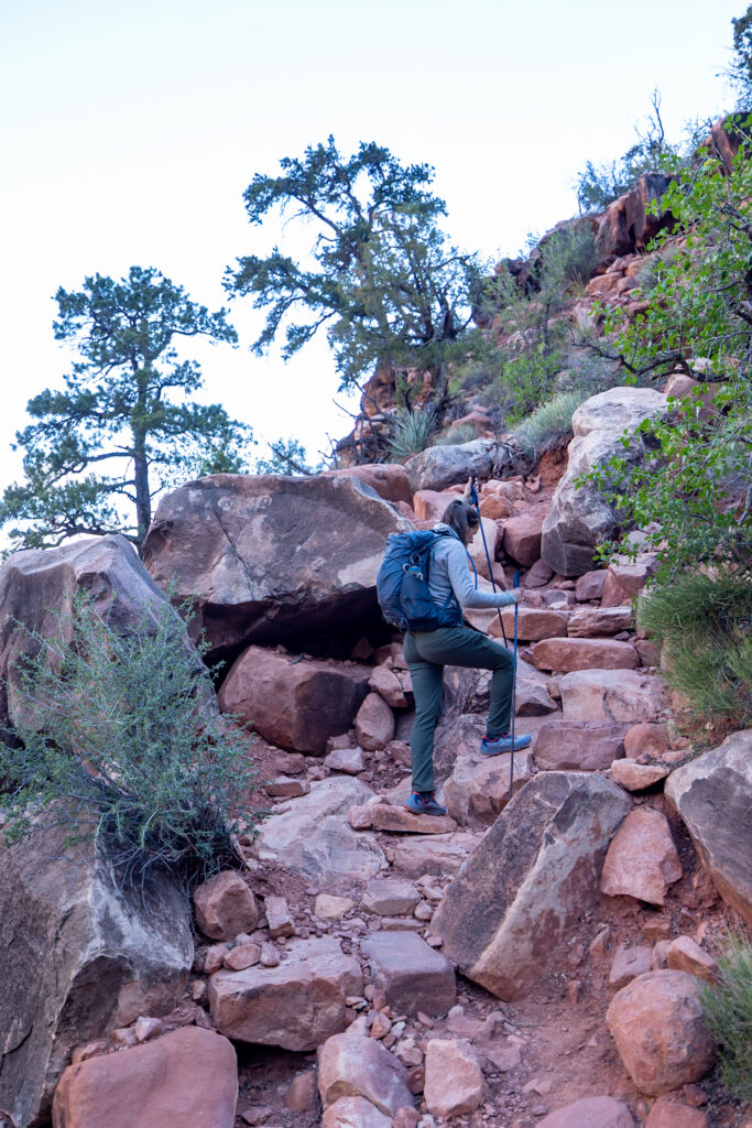 Jen hiking up a steep rocky trail with red stone formations and scattered vegetation on the Grand View Trail