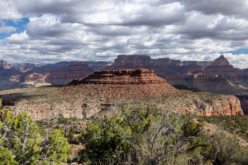 Expansive view of the Grand Canyon from Grand View Trail showing layered red rock formations, desert vegetation in the foreground, and cloudy sky above