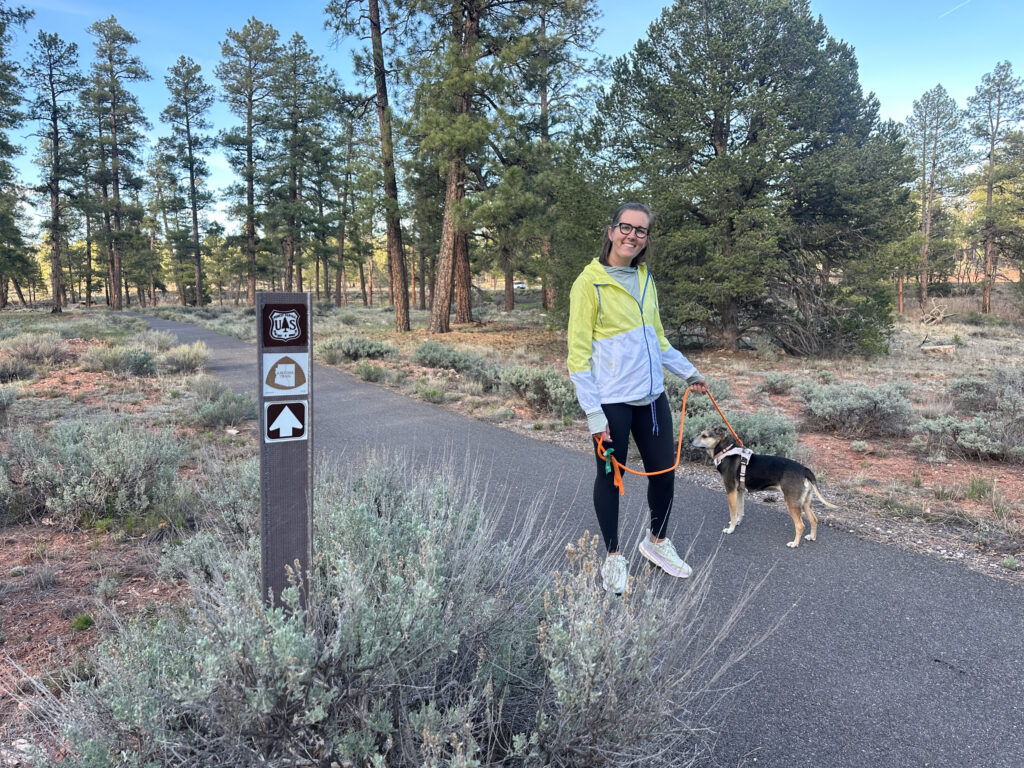 Hiker with dog on paved Greenway Trail surrounded by ponderosa pines and desert shrubs, with trail marker visible