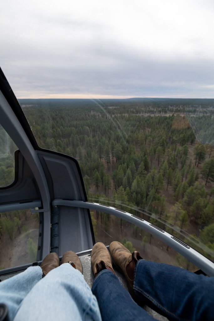 View from inside a helicopter showing two people's feet dangling over a vast forested landscape with canyon terrain visible below