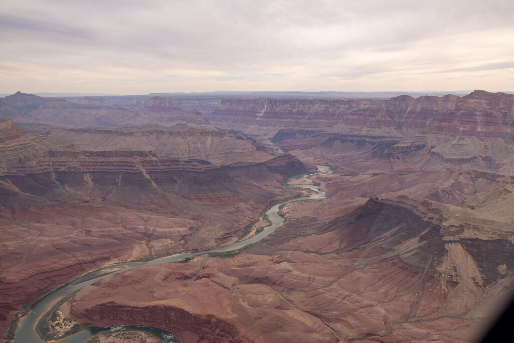 Aerial view of the Grand Canyon showing layered red rock formations, the Colorado River winding through the canyon, and the North Rim in the distance, taken during a helicopter tour