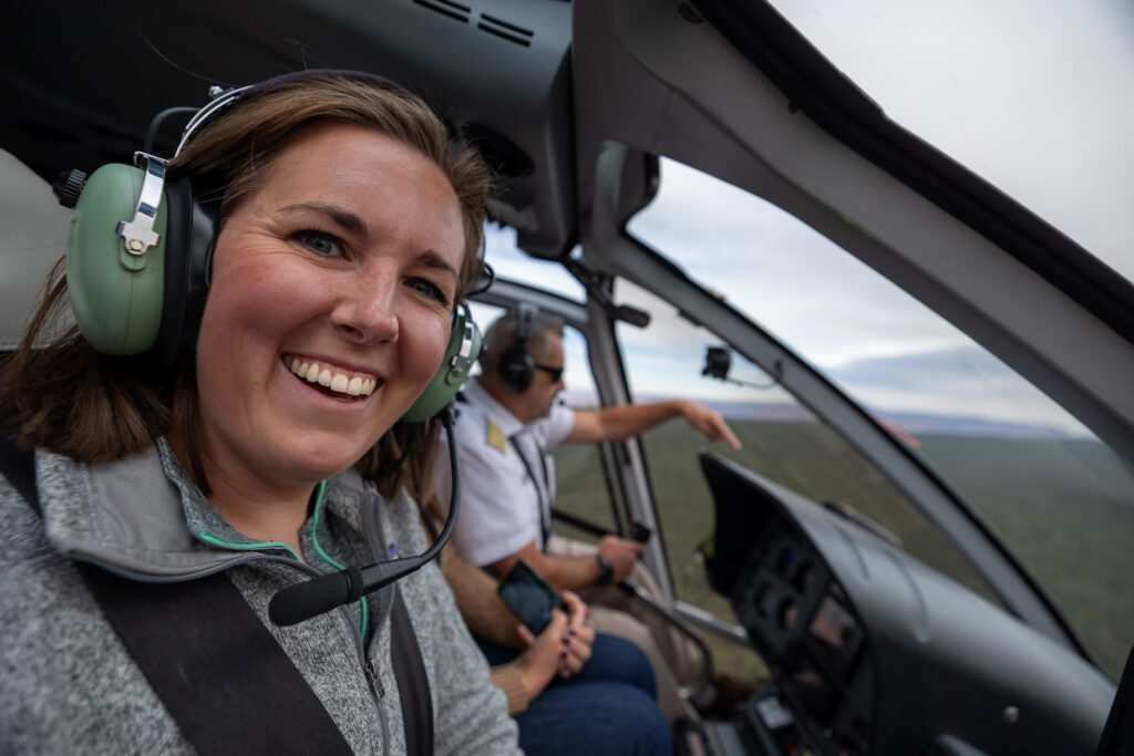 Jen smiling while wearing headphones inside a helicopter during a Grand Canyon tour, with pilot and landscape visible through the window