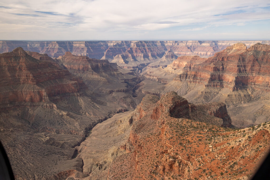 Aerial view of the Grand Canyon showing layered red rock formations, the Colorado River winding through the canyon floor, and expansive vistas of both the North and South Rims under a partly cloudy sky