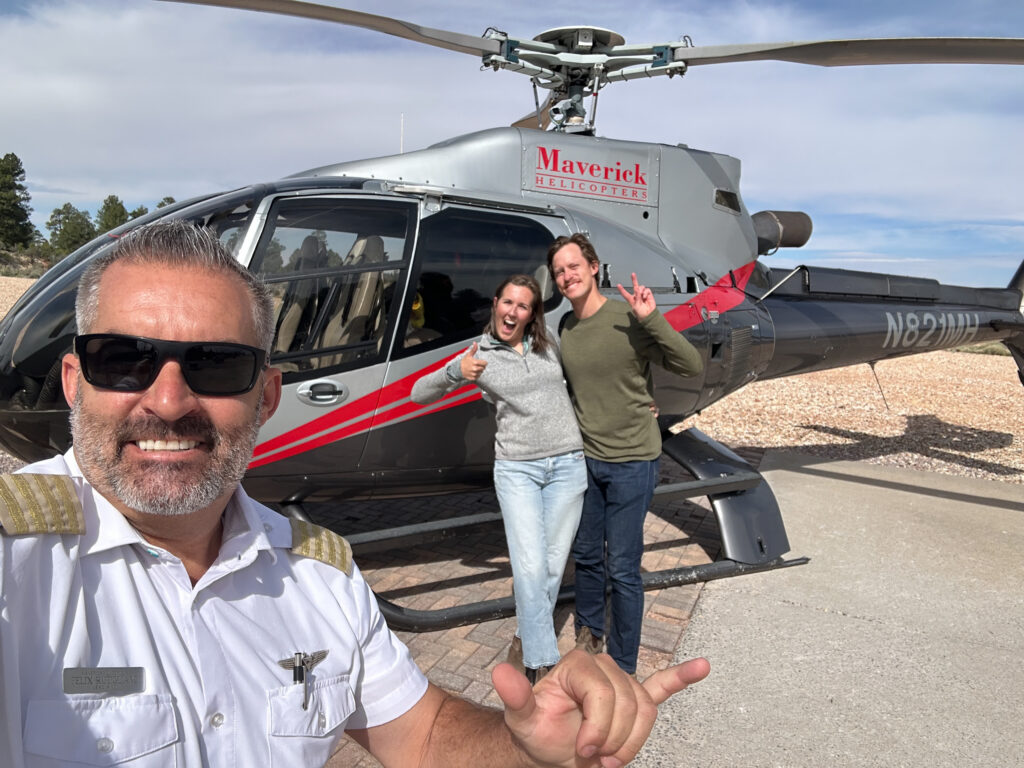 Helicopter captain in white uniform and sunglasses posing with two passengers in front of a Maverick Helicopters aircraft on the ground