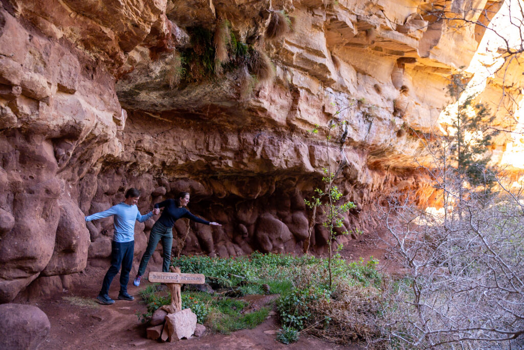 Two hikers standing at the entrance to a large rock shelter with a wooden sign reading "Dripping Springs" beneath a towering red sandstone cliff face