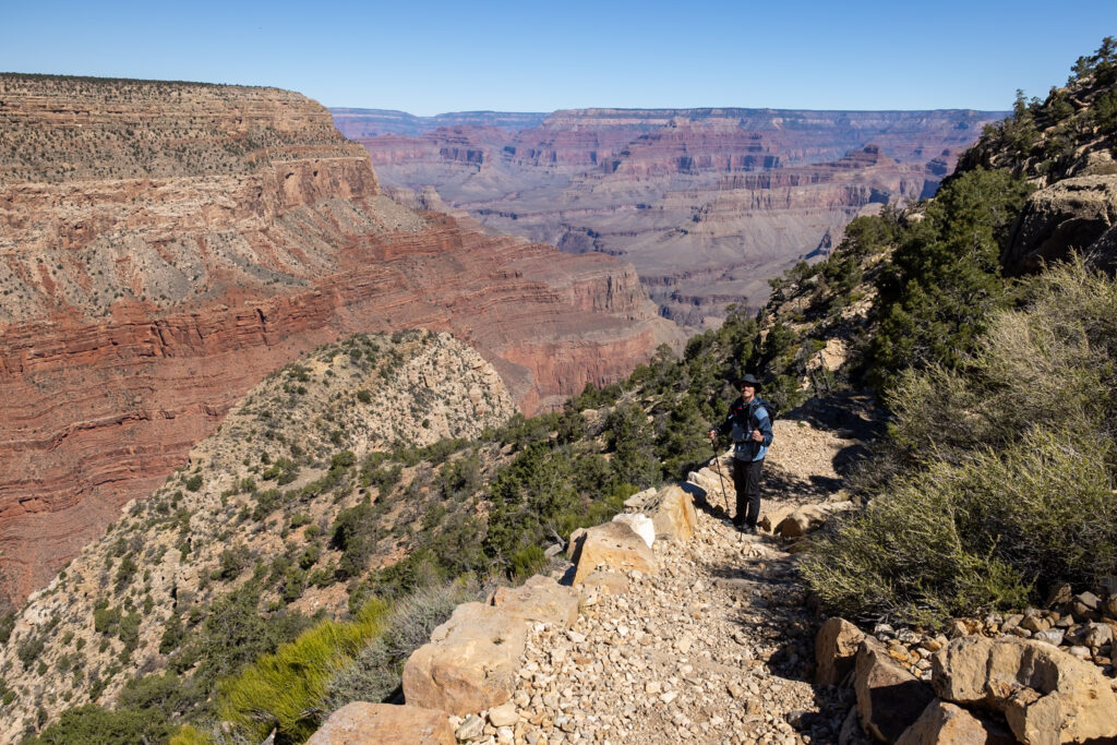 Hiker descending rocky trail through desert vegetation with expansive canyon views and layered red rock formations in the background