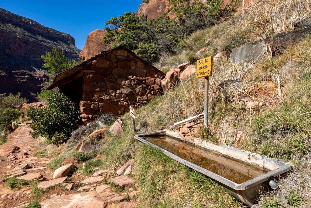 Santa Maria Springs water source on the Hermit Trail, featuring a stone shelter, yellow sign, and metal trough with water, surrounded by canyon vegetation and red rock formations