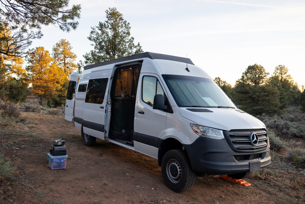 White Mercedes van with open side door parked on dirt ground surrounded by pine and aspen trees in Kaibab National Forest near Grand Canyon