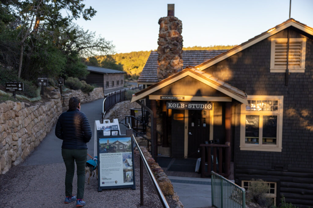 Woman walking on a path toward Kolb Studio, a historic stone building with signage, in Grand Canyon Village at sunset. This is one of the best things to do on the Grand Canyon South Rim.