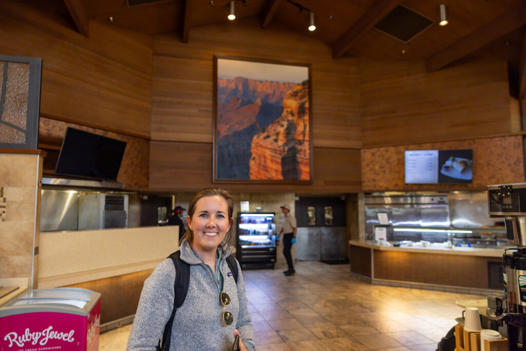 Woman smiling in the Maswik Food Court, a modern dining facility with wood paneling, service counters, and a Grand Canyon photograph on the wall