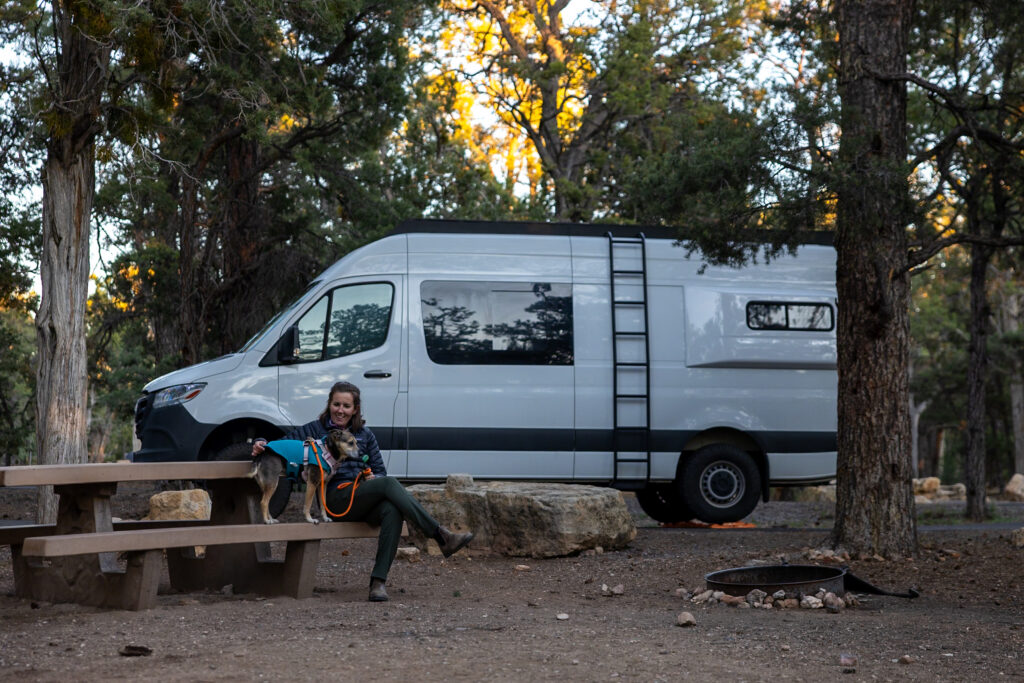 Jen and Ollie relaxing at their campsite at Mather Campground, with a white camper van and tall pine trees in the background