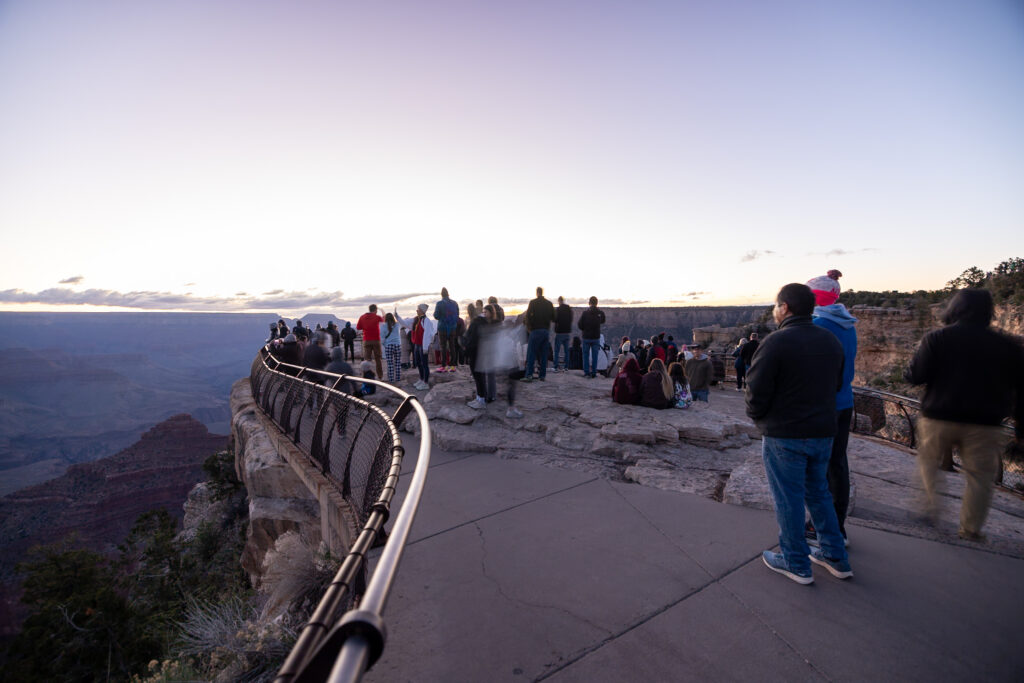Large crowd of visitors gathered at Mather Point viewpoint during sunrise, with the Grand Canyon visible in the background and early morning light on the horizon
