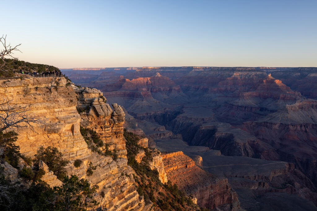Golden morning light illuminates the layered rock formations and canyon walls of the Grand Canyon at sunrise, with visitors visible on the rim at left