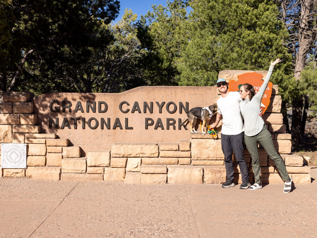 Two people and a dog posing at the Grand Canyon National Park entrance sign, one with arm raised, surrounded by pine trees