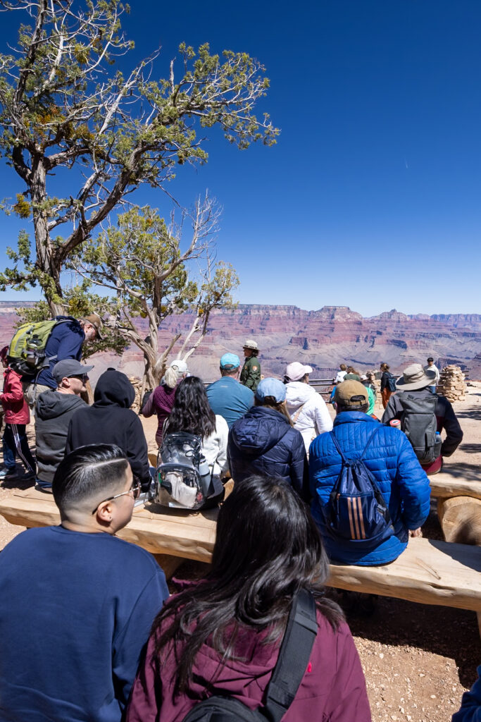 Group of visitors gathered at an overlook listening to a park ranger present near the Geology Museum, with the Grand Canyon visible in the background. This is one of the best things to do at the South Rim at the Grand Canyon.
