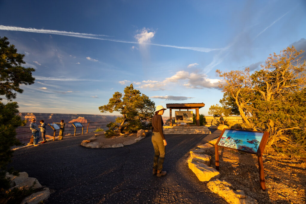 Jen with a sun hat standing on the Rim Trail at Pima Point during golden hour, with the Grand Canyon and other visitors visible in the background
