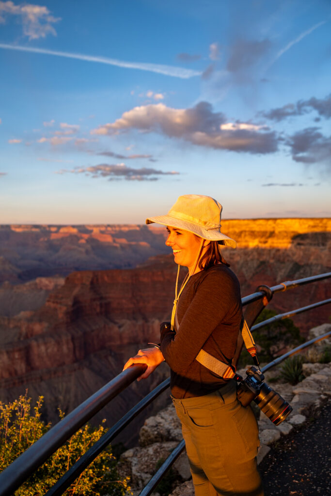 Jen in yellow tan and black shirt standing at a railing overlooking the Grand Canyon at sunset, with golden light illuminating the canyon layers and sky filled with clouds