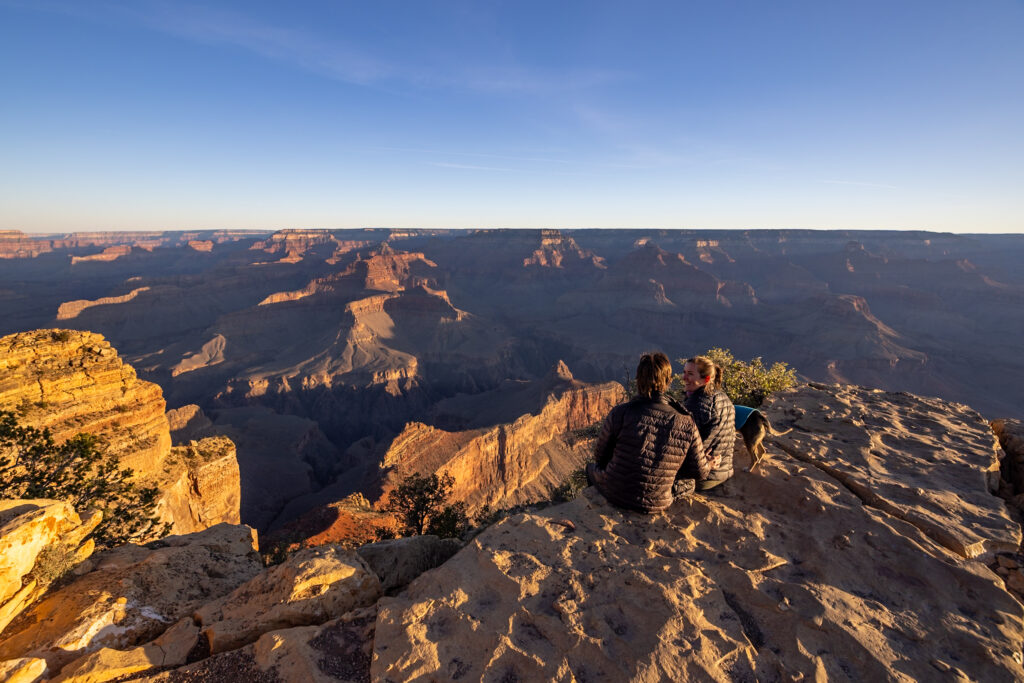 Two hikers sitting on the rim overlooking the Grand Canyon's layered rock formations and deep valleys under clear blue sky at sunset