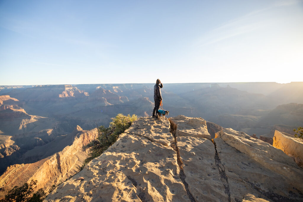 Elliot and Ollie (a dog) standing on a rocky outcrop at Powell Point overlooking the Grand Canyon at sunrise, with golden light illuminating the canyon walls and the Colorado River below