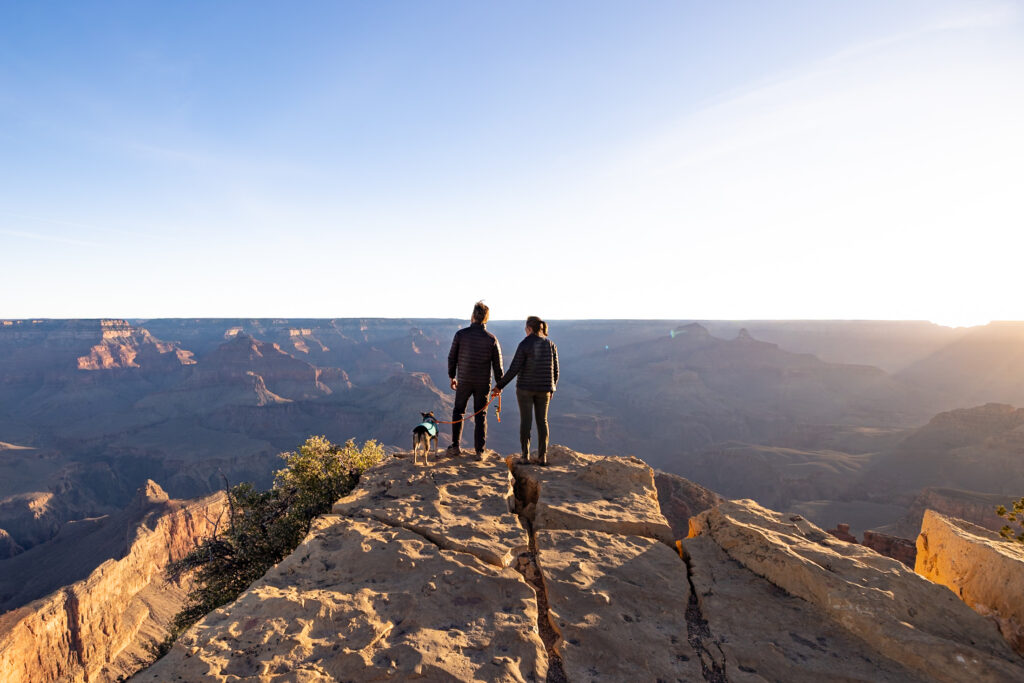 Two hikers and a dog standing on a rocky outcrop at Powell Point, overlooking the vast expanse of the Grand Canyon under clear blue sky