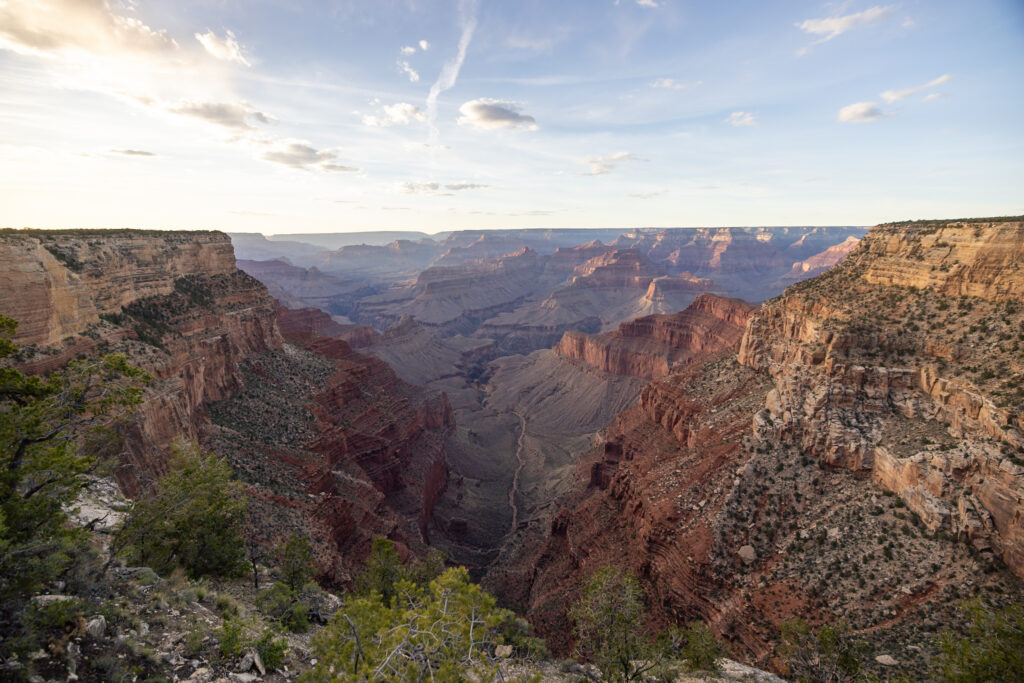 Expansive view of the Grand Canyon showing layered red rock formations, the Colorado River winding through the canyon floor, and desert vegetation on the rim under a partly cloudy sky