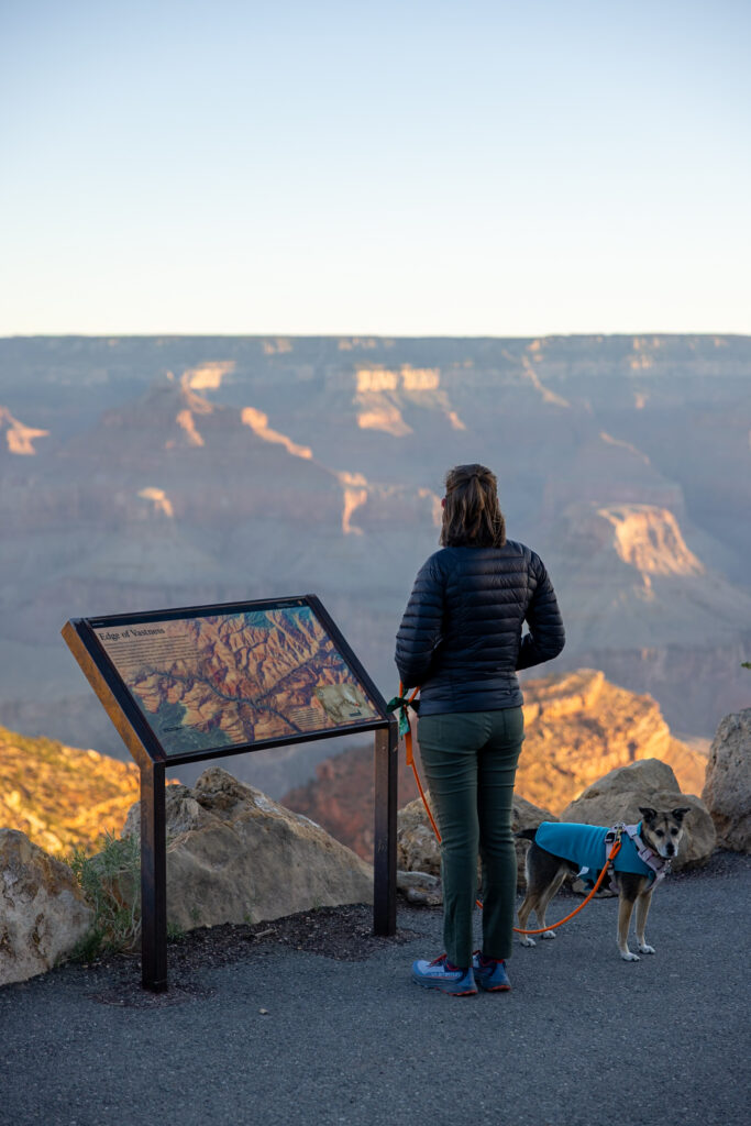 Woman and dog standing at an interpretive sign on the Rim Trail, overlooking the Grand Canyon's layered rock formations in golden light