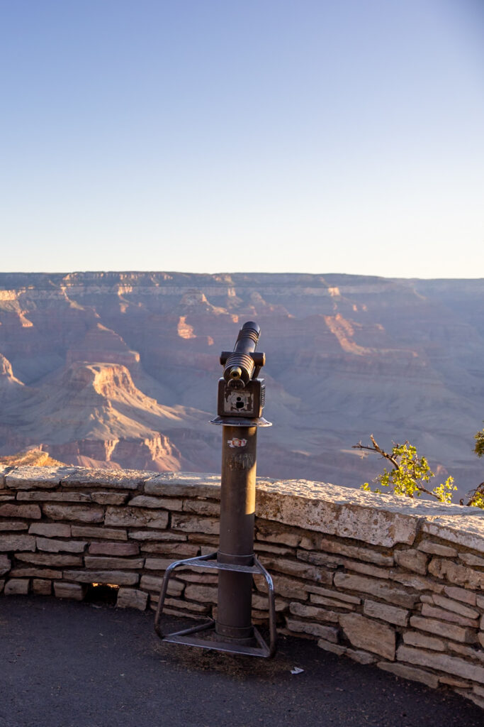 Coin-operated telescope on a stone wall overlooking the Grand Canyon's layered rock formations at sunset