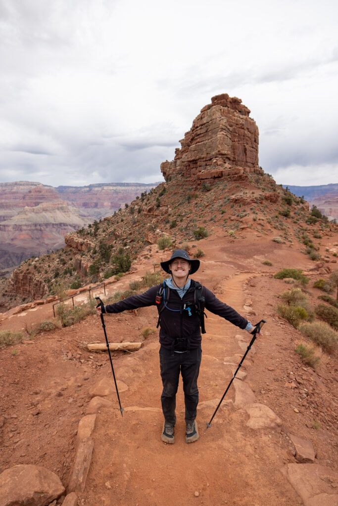 Hiker on South Kaibab Trail with trekking poles, standing on red dirt path with distinctive rock formation and canyon landscape behind